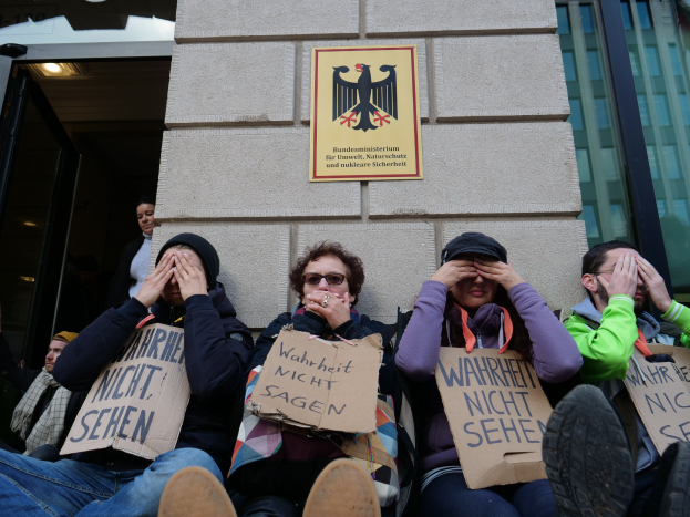Eine Gruppe von Menschen sitzt vor einem Gebäude, hält Schilder hoch, einige bedecken ihre Gesichter, mit einem Logo und einer Tafel an der Wand und einer Glastür auf der rechten Seite.