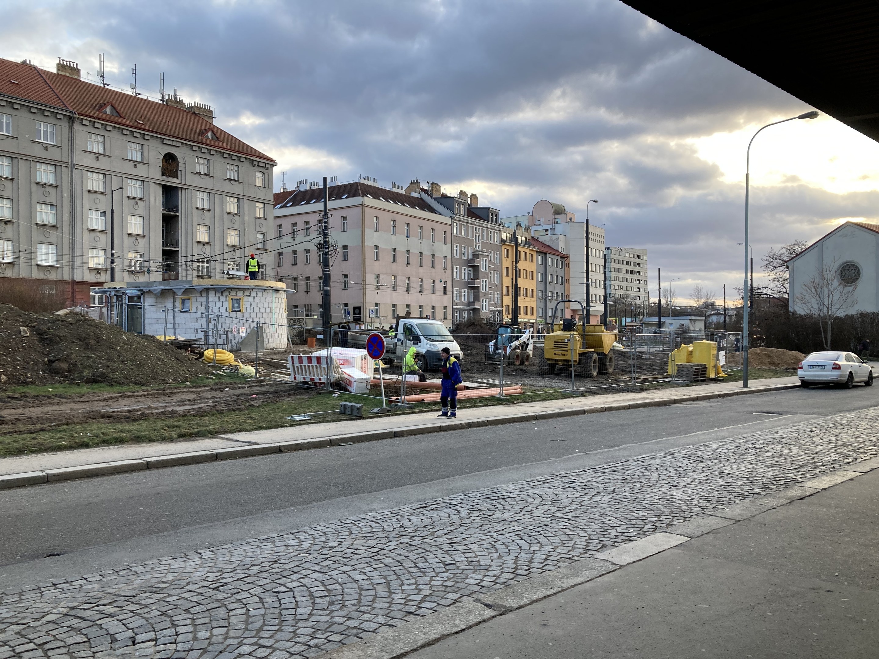 Eine Stadtstraße mit parkenden Autos, Gebäuden, Laternen, Bäumen und einem bewölkten Himmel, mit einer Straßensperre mit Fahrzeugen und Fußgängern im Vordergrund.