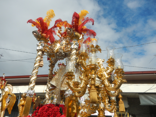 Ein großer goldener und roter Festwagen, der mit Blumen und anderen Dekorationsgegenständen geschmückt ist, in einem Karnevalsumzug mit einem Gebäude, Strommasten mit Drähten und einem bewölkten Himmel im Hintergrund.