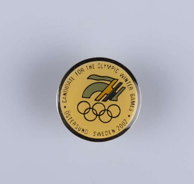 Ein runder Abzeichenaufkleber mit schwarzem Hintergrund und goldfarbenem Olympia-Winterspiellogo in der Mitte, mit "Olympic Winter Games" in fetter weißer Schrift.