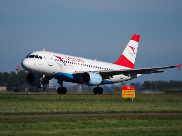 Ein Airbus A320-200 der Austrian Airlines startet vom Frankfurter Flughafen, mit einer Tafel im Vordergrund, Gras, Bäumen, Gebäuden und einem klaren blauen Himmel im Hintergrund.