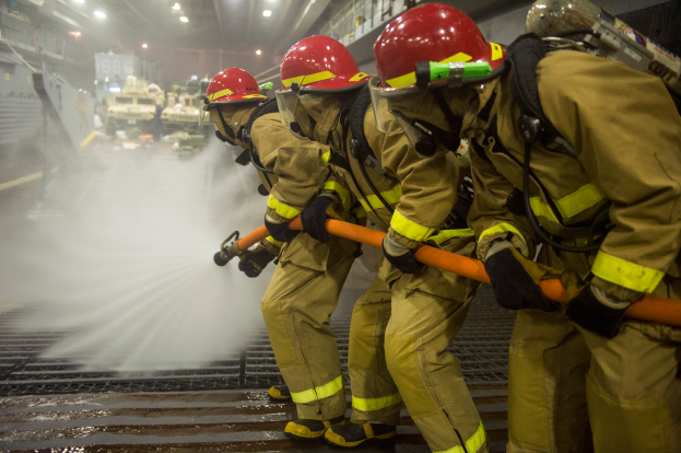 Eine Gruppe von Feuerwehrmännern in Helmen und Handschuhen hält Schläuche und spritzt Wasser auf ein Feuerwehrauto, mit verschiedenen Gegenständen und Texttafeln im Hintergrund und einem Boden unten.