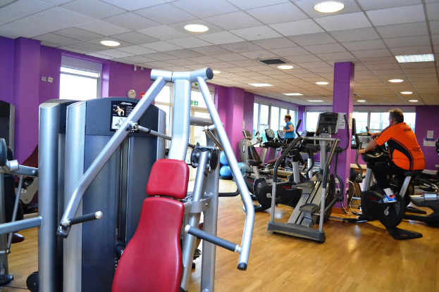 Ein Fitnessstudio mit verschiedenen Geräten und einigen Menschen beim Training, mit einer Decke und Lampen oben sichtbar.
