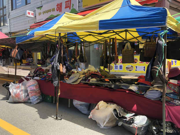 Ein Markt mit Zelten, die Taschen, Hüte und andere Gegenstände präsentieren, vor Häusern mit Fenstern und Texttafeln.