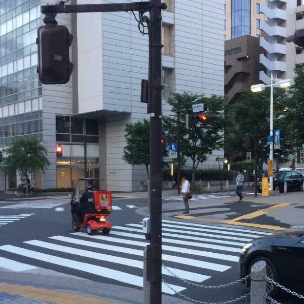 Ein Scooter auf einer Straße mit Menschen im Hintergrund, eine Ampel im Vordergrund und Gebäude mit Bäumen im Hintergrund.