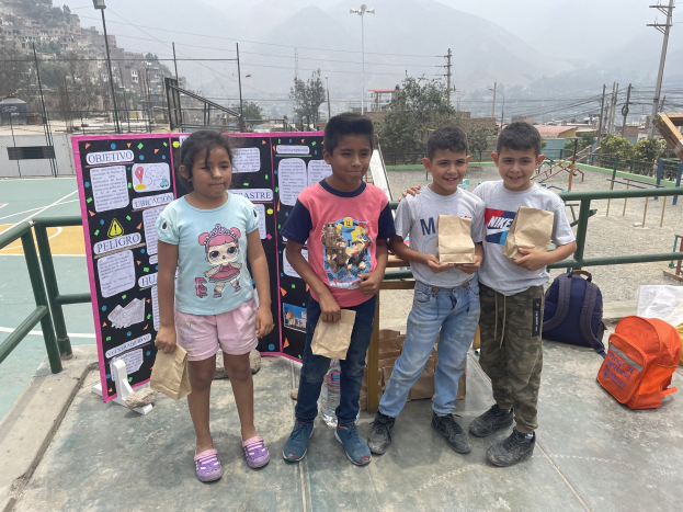 Gruppe von Kindern auf einem Basketballplatz mit Papiertüten in den H├Ąnden, mit Tüten und Gegenst├Ąnden rechts und einer Tafel mit Text links, vor dem Hintergrund von Z├Ąunen, Masten, Stra├čenlaternen, B├Ąumen, Geb├Ąuden, H├Ąngen und Himmel.