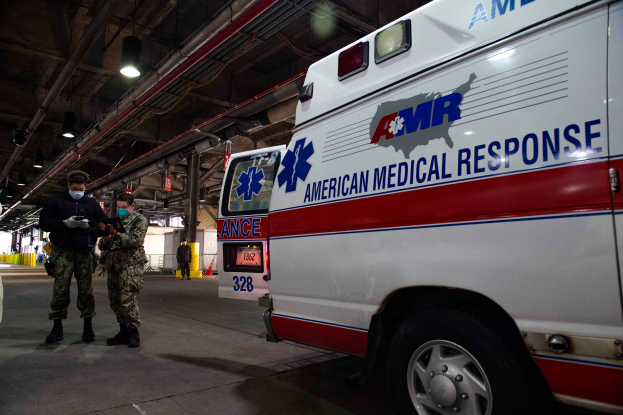 Ein Rettungswagen der American Medical Response (AMR) steht in einem Gebäude, vor ihm stehen zwei maskierte Personen, links ein Fahrzeug und im Hintergrund mehrere Personen, Lampen und Rohre.