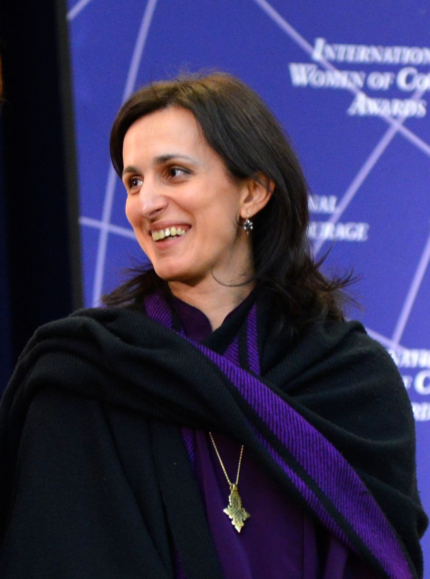 Eine Frau in einem lila Kleid und schwarzer Stola steht neben einer anderen lachenden Frau, mit einem Banner im Hintergrund, auf dem 'Internationale Frauen der Farbe Awards' steht.