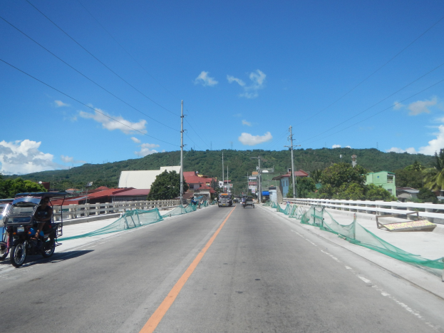 Autobahn mit fahrenden Fahrzeugen, ein geparktes Motorrad auf dem Seitenstreifen, Geländer, Strommasten mit Drähten, Bäume, Gebäude, entfernte Hügel und ein bewölkter Himmel.
