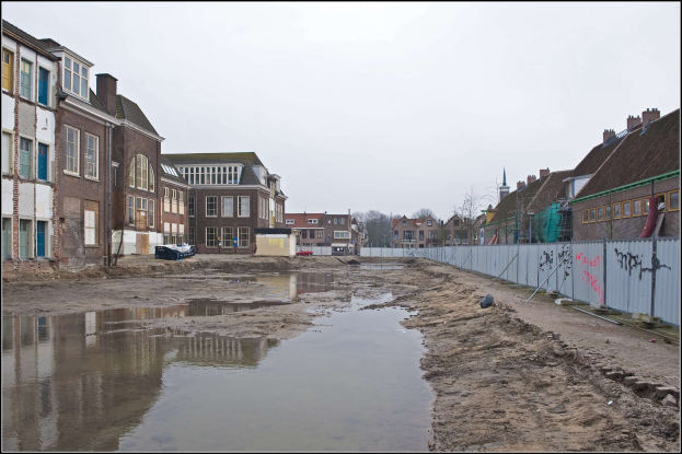 Flutstraße in der Stadtmitte mit Wasser auf dem Boden, ein Zaun auf der rechten Seite, Gebäude mit Fenstern auf der linken Seite, Bäume im Hintergrund und Himmel darüber.