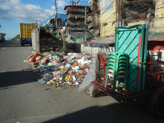 Ein Lastwagen neben einem Müllhaufen auf einer Straße, mit einem mit Plastikstühlen beladenen Handwagen rechts und Gebäuden, Strommasten, Bäumen und einem bewölkten Himmel im Hintergrund.