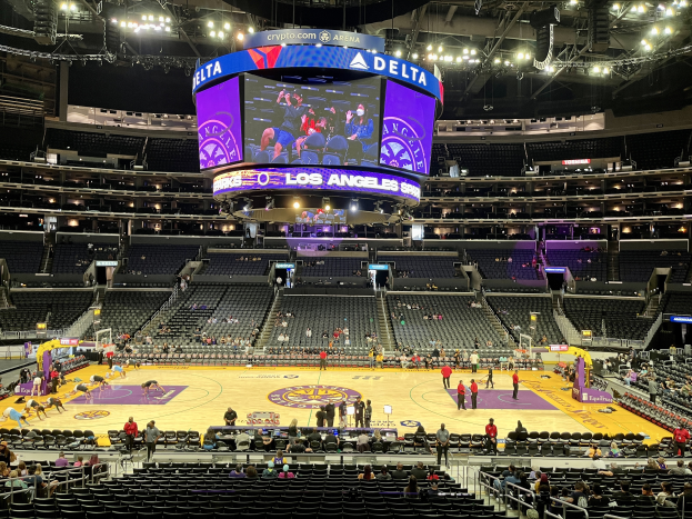 Eine große Arena voller Zuschauer, die ein Basketballspiel verfolgen, mit einem großen Bildschirm und Scheinwerfern, die den Court beleuchten.