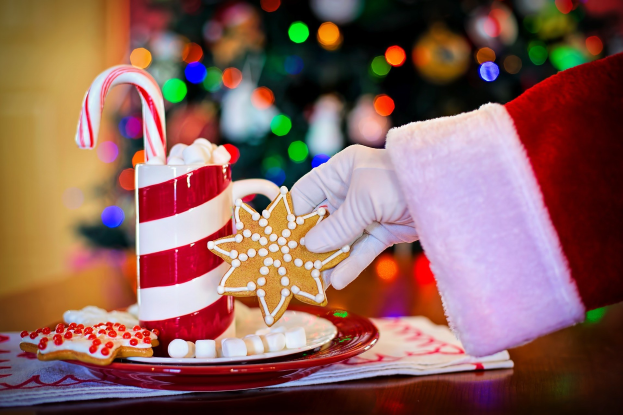 Eine Person mit einem Santa Claus Hut, die einen Becher heißen Kakao und einen Bonbonstab hält und ein Teller mit Keksen und ein Tuch auf dem Tisch vor ihnen steht, vor einem Weihnachtsbaum und einer Wand.