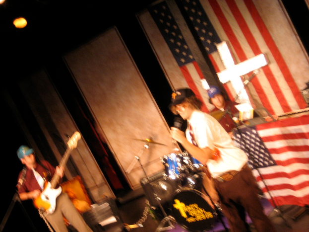 Eine Gruppe von Menschen, die Musikinstrumente mit zusätzlicher Ausrüstung spielen, mit einer amerikanischen Flagge im Hintergrund und einer Wand im Hintergrund.