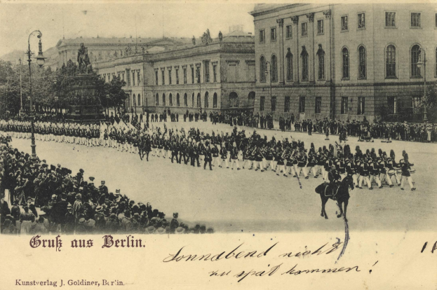 Ein altes Schwarz-Weiß-Foto einer Parade in Berlin, Deutschland, mit Menschen auf dem Boden und auf Pferden, einer Statue, Straßenmästen, Straßenlaternen, Bäumen, Gebäuden mit Fenstern und einem bewölktem Himmel, mit Text unten.