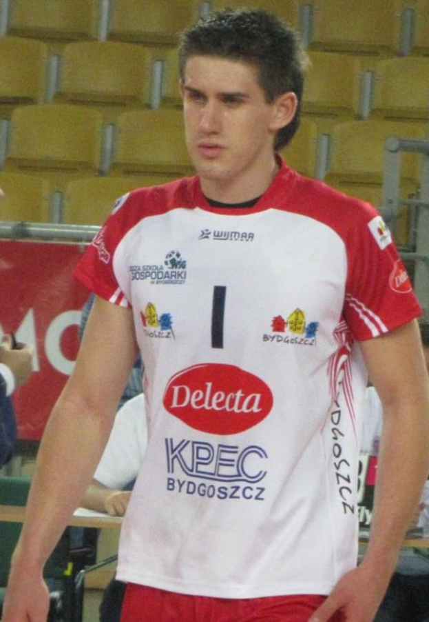 Ein Mann in einem roten und weißen Handballtrikot steht auf einem Feld, entschlossen, mit Zuschauern und einer roten Fahne mit der Aufschrift "Krasnoyarsk vs Bydgoszcz" im Hintergrund.
