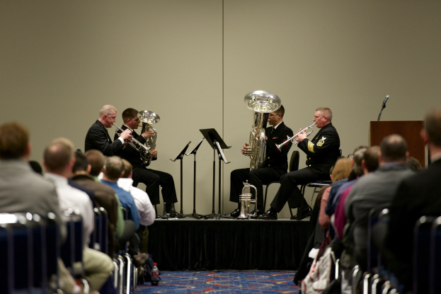 Eine Gruppe von Männern, die Brass-Instrumente auf einer Bühne spielen, mit Notenständern, einem Mikrofonpodest und einem Publikum, das auf Stühlen sitzt.