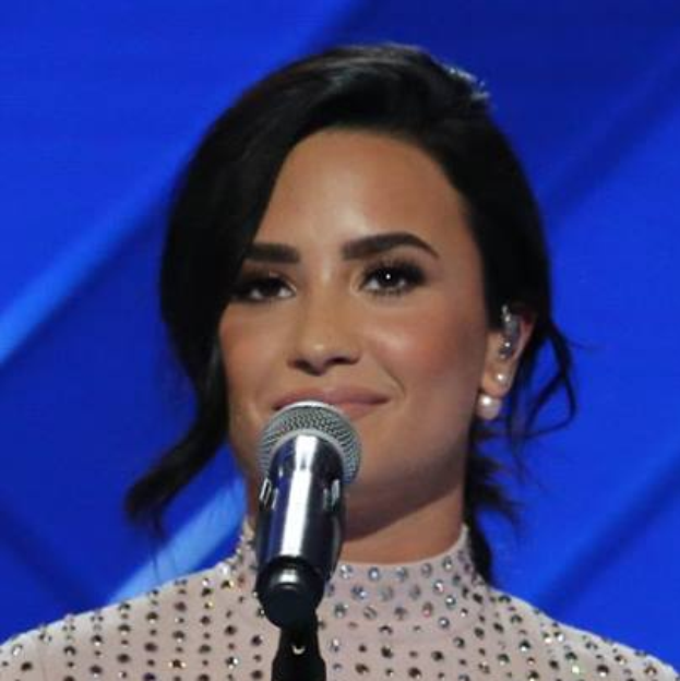 Demi Lovato in einem schwarzen Kleid mit einem sleek Bob vor einem Mikrofon an den Grammys posend, mit einem blauen Hintergrund.