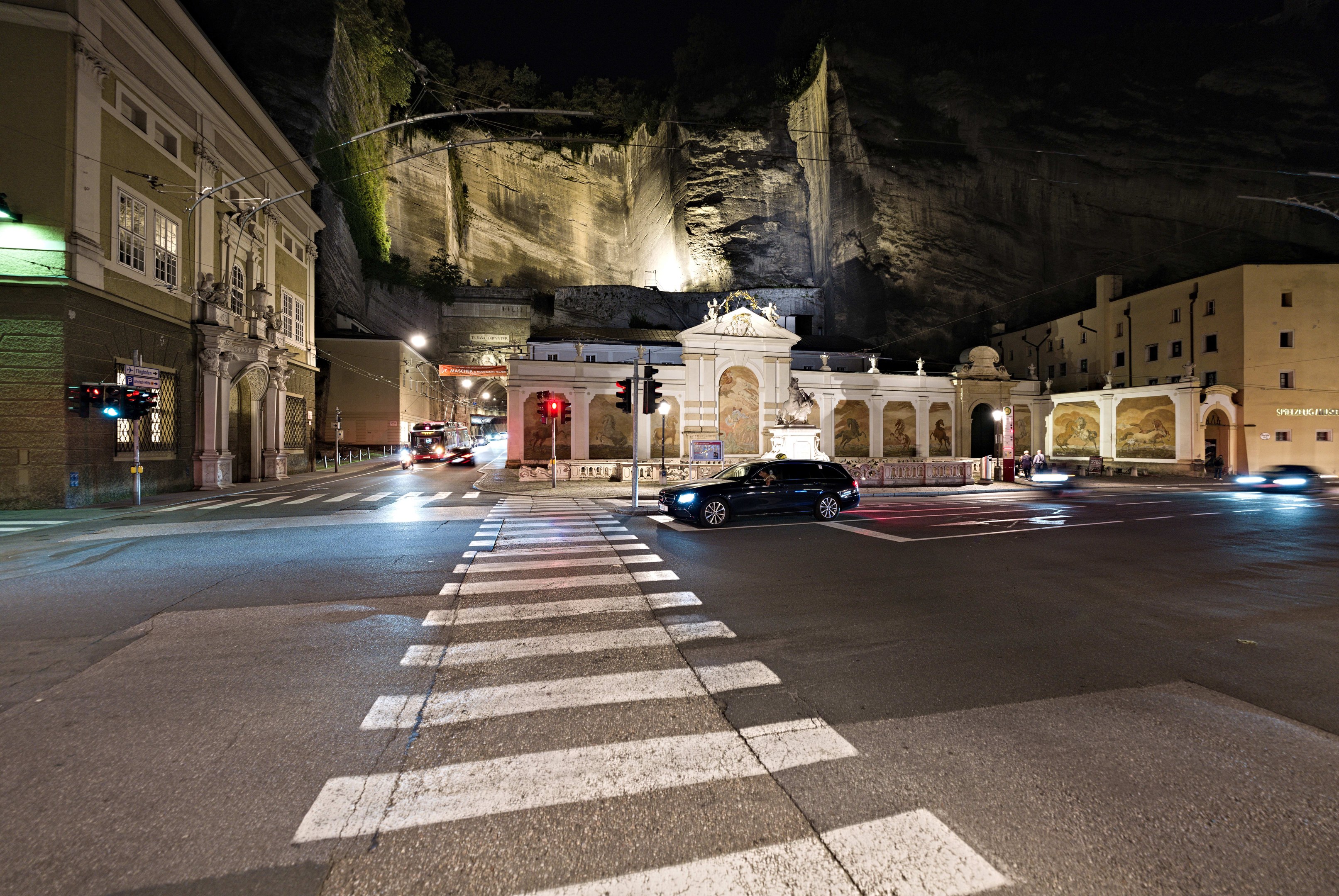 Eine nächtliche Stadtstraße mit fahrenden Autos, Straßenlaternen, Ampeln, beleuchteten Gebäuden, Laternenpfählen, einem Hügel im Hintergrund und einer Zebrastreifenfurt im Vordergrund.