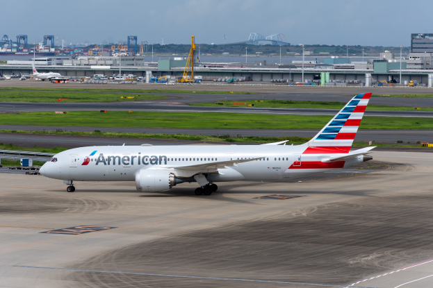 American Airlines-Flugzeug auf der Rollbahn eines Flughafens mit Gras, Fahrzeugen, Gebäuden, Masten, Kränen, Bäumen und einem klaren blauen Himmel im Hintergrund.