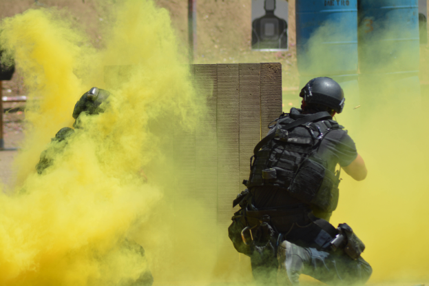 Zwei Polizisten in Einsatzausrüstung mit gelbem Rauch aus dem Mund, mit Helmen und Schutzausrüstung vor einer Wand mit einer Tafel und anderen Gegenständen, mit sichtbarem Boden.