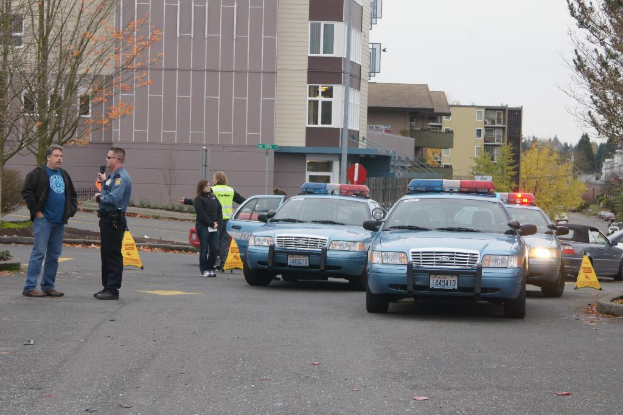 Autos auf einer Straße mit vier Menschen in der Nähe, Gebäude mit Fenstern im Hintergrund, Bäume und Warn Dreiecke.