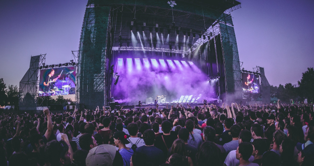 Eine große Menschenmenge bei einem Musikfestival schaut auf einer hell erleuchteten Bühne mit Instrumenten und Bildschirmen, die vor einem Gebäude, Bäumen und einem klaren blauen Himmel stehen.