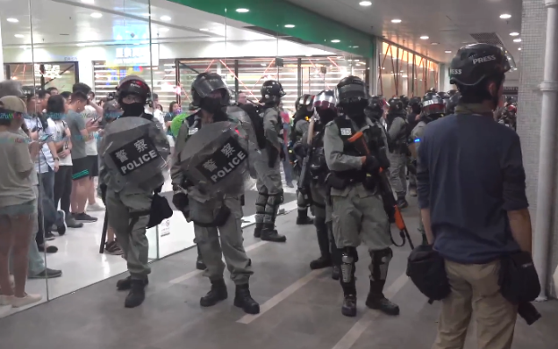 Eine Gruppe von Polizeibeamten steht vor einer Menschenmenge, einige tragen Helme und halten Waffen, in einem Einkaufszentrum mit einer Säule auf der rechten Seite, einer Glaswand im Hintergrund und Lampen an der Decke.