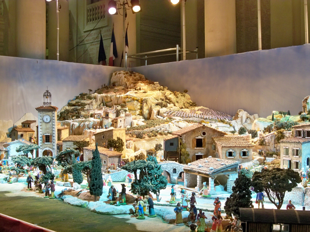 Eine Museumsausstellung eines Krippenmodells mit Menschen, Häusern, Bäumen, Steinen, einem Uhrturm, Fahnen, Pfählen und Lichtern, alles beleuchtet von Museumsbeleuchtung.