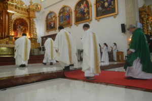 Eine Gruppe von Priestern geht über einen roten Teppich in einer Kirche, während Menschen vor und rechts von ihnen auf dem Boden stehen und Foto Rahmen an der Hintergrundwand hängen.
