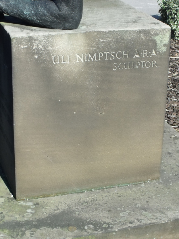 Ein Stein mit Text in der Mitte, möglicherweise eine Statue oben, trockene Zweige, Erde und Pflanzen rechts, mit einer Straße im Hintergrund.