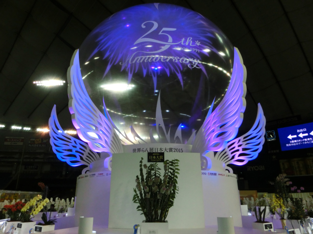 Ein großer Kristallball mit Flügeln oben drauf, umgeben von Blumensträußen, Namensschildern und anderen Gegenständen auf dem Boden, vor einem festlichen Hintergrund mit Schildern, Lichtern und einem Bildschirm für ein 25-jähriges Jubiläum.