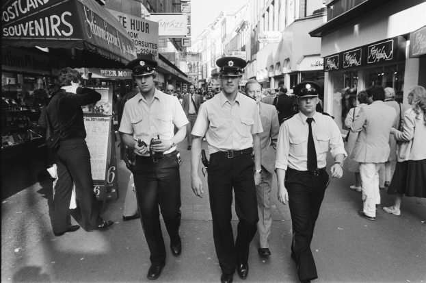 Drei Polizisten in Uniform gehen eine belebte Straße mit hohen Gebäuden und Geschäften entlang, mit Passanten und einem sichtbaren Himmel im Hintergrund.