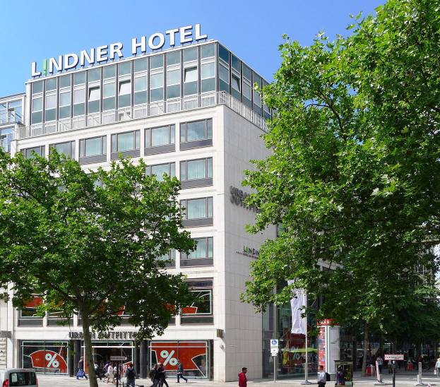 Ein großes modernes Hotelgebäude mit Glasfenstern und Beschriftung oben, umgeben von Bäumen und einer belebten Straße mit Fußgängern und Fahrzeugen unter einem sichtbaren Himmel.