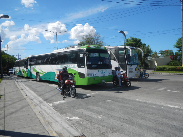 Ein grüner und weißer Shuttlebus steht am Straßenrand mit Motorradfahrern davor, ein grasbewachsener Fußweg links daneben und Gebäude, Bäume und Laternen im Hintergrund unter einem klaren blauen Himmel.