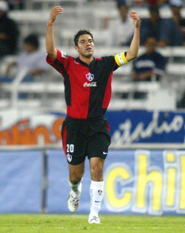 Ein Fussballspieler in rot-schwarzem Trikot rennt mit ausgebreiteten Armen auf einem Feld, umgeben von einer Menge im Publikum, mit einem "Chivas de Guadalajara"-Schild im Hintergrund.