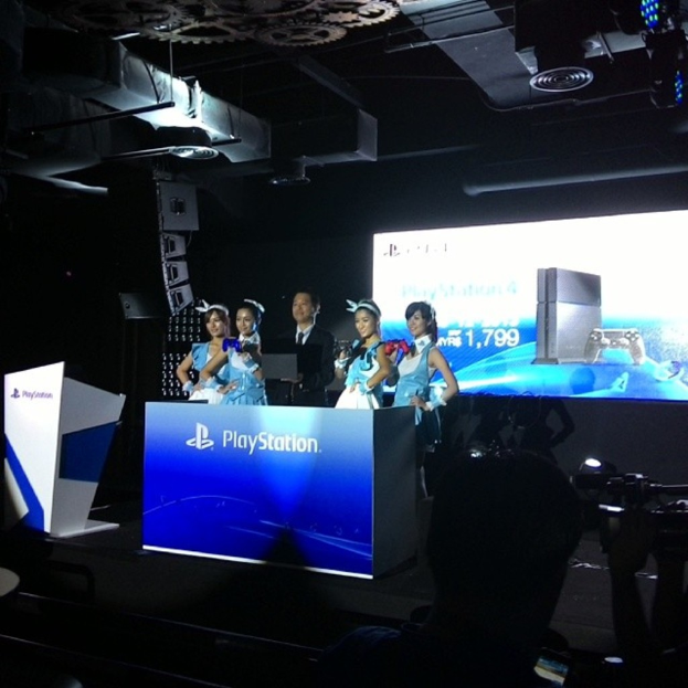 Menschen auf einer Bühne neben einem Podium mit "Playstation" darauf, mit einem Projektionsschirm an der Wand dahinter.