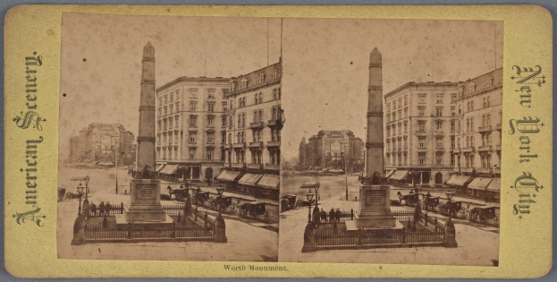 Altes Schwarz-Weiß-Foto einer Stadtstraße mit einem zentralen Denkmal, umgeben von Gebäuden, Straßenmöbeln, parkenden und fahrenden Fahrzeugen, Fußgängern auf dem Gehweg und einem sichtbaren Himmel.