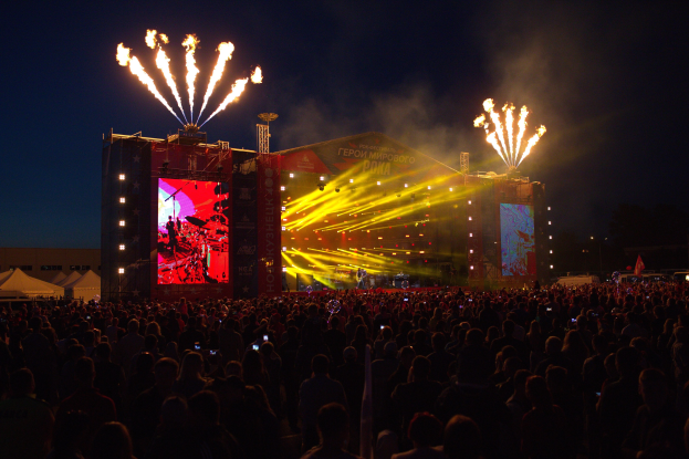 Eine große Menge Menschen bei einem Musikfestival schaut auf eine hell erleuchtete Bühne mit Zelten links daneben und Feuerwerk am dunklen Himmel dahinter.