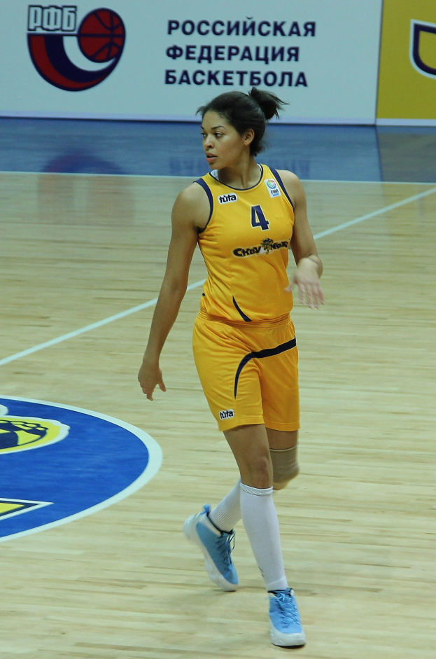 Ein professioneller Basketballspieler in einem gelben Trikot und Sportschuhen dribbelt auf einem Court mit einer Tafel im Hintergrund.