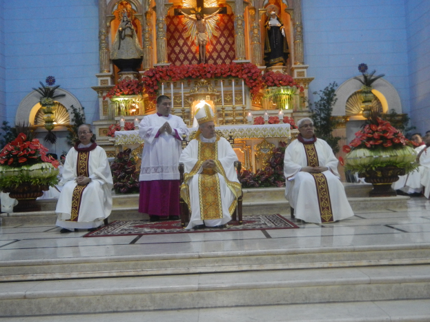 Eine Gruppe von Priestern sitzt auf den Stufen einer Kirche, umgeben von einem Teppichboden, Blumensträußen, Statuen, Kerzen mit Ständern und anderen Gegenständen, wobei die Kirchenwände im Hintergrund sichtbar sind.