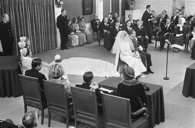 Schwarzes und weißes Foto einer Hochzeitszeremonie mit sitzenden und stehenden Gästen, einem Mann mit Mikrofon, einer Frau mit Blumenstrauß, einem Podium mit Mikrofon, Blumengestecken, einem Vorhang, einem Rahmenfoto an der Wand und einer Tür im Hintergrund.