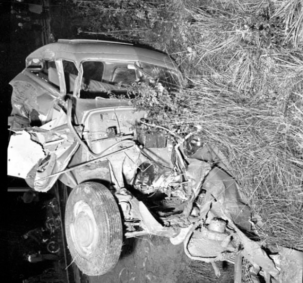 Ein Schwarz-Weiß-Foto eines schwer beschädigten Autos am Straßenrand, mit eingedellter Front und zersplitterten Fenstern, einem Baum im Hintergrund und verstreuten Gegenständen an der Szene.