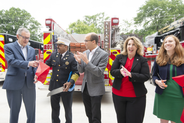 Gruppe von Menschen bei einer Eröffnungszeremonie für die Alexandria Fire Department, die vor einem Feuerwehrauto klatschen und lächeln, vor einem Hintergrund mit Bäumen und einem klaren blauen Himmel.