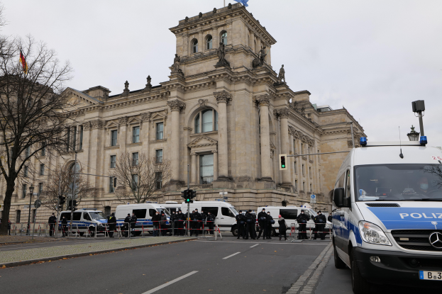 Eine Gruppe von Polizeibeamten steht vor dem Reichstaggebäude in Berlin, Deutschland, mit Fahrzeugen, einem Zaun, Verkehrsampeln, Laternen, Bäumen und Fahnen im Hintergrund, bei klarem Himmel.