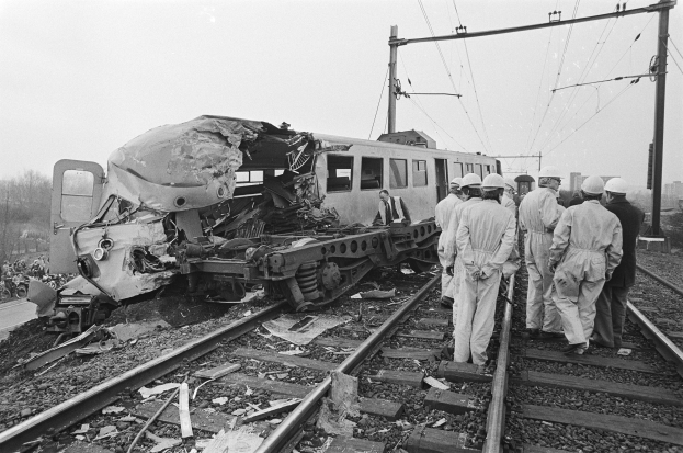 Eine Schwarz-Weiß-Szene eines Zugunglücks mit Menschen in Helmen drumherum, Strommasten, Bäumen, Gebäuden und einem klaren Himmel im Hintergrund.