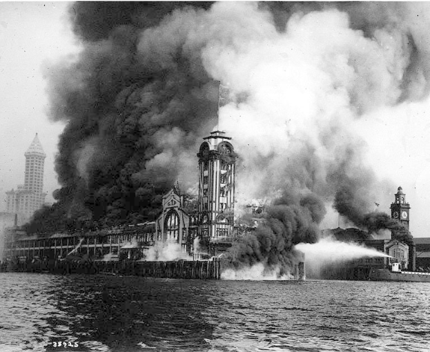 Schwarz-weißes Foto eines großen, von Flammen umloderten Gebäudes mit Rauch, der herausquillt, einem Gewässer im Vordergrund und anderen Gebäuden und Himmel im Hintergrund.