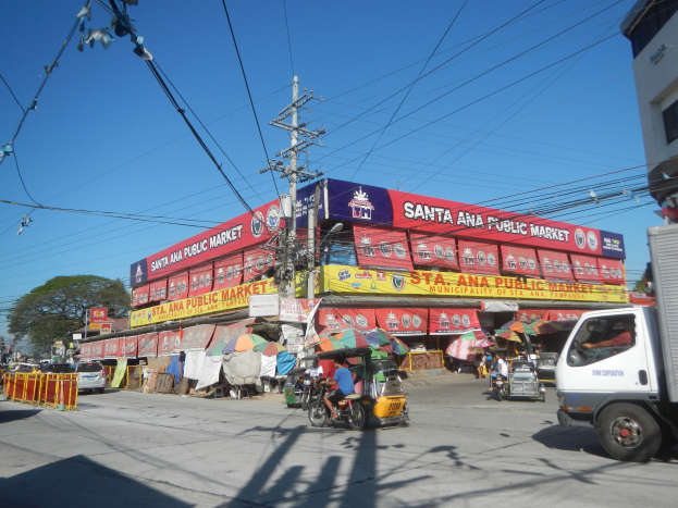 Eine belebte Straßenszene mit einem Lastwagen, mehreren Fahrzeugen, Fußgängern, einem Strommast mit Drähten, Bäumen und einem klaren blauen Himmel.
