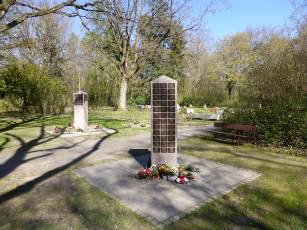 Kriegerdenkmal auf einer grünen Wiese mit Blumensträußen, einer Bank, Bäumen, Gräbern und einem klaren blauen Himmel.