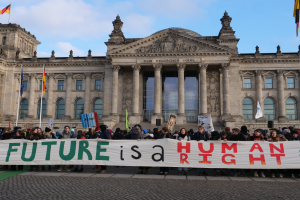 Eine Gruppe von Menschen hält ein Transparent mit der Aufschrift "Zukunft ist ein Menschenrecht" vor dem Reichstagsgebäude in Berlin, Deutschland, das mit Fenstern, Säulen, Bögen und Flaggenmasten geschmückt ist und von einer bewölkten Himmel überlagert wird.